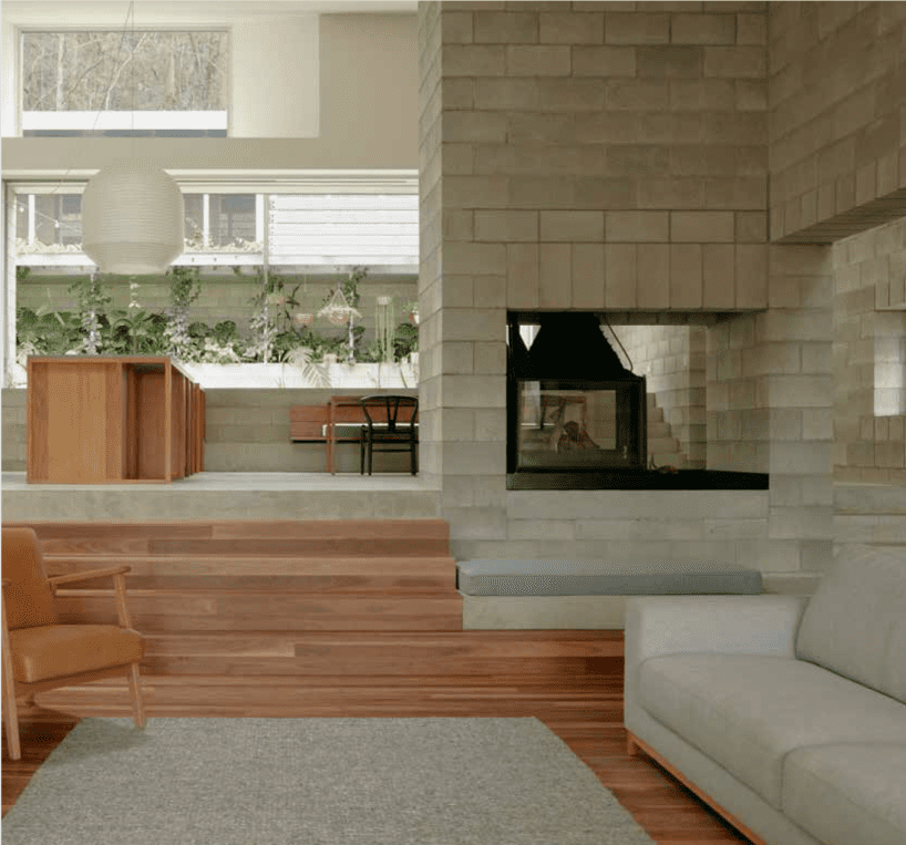 A modern living room features wooden floors and an Austral Bricks fireplace. A gray sofa sits on the right, while a brown armchair is on the left. The room steps up to a dining area adorned with a large pendant light and plants on the window ledge.